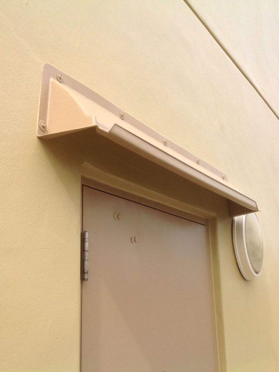 A beige door rain guards mounted above a single hollow metal door with wall mount light to the right.
