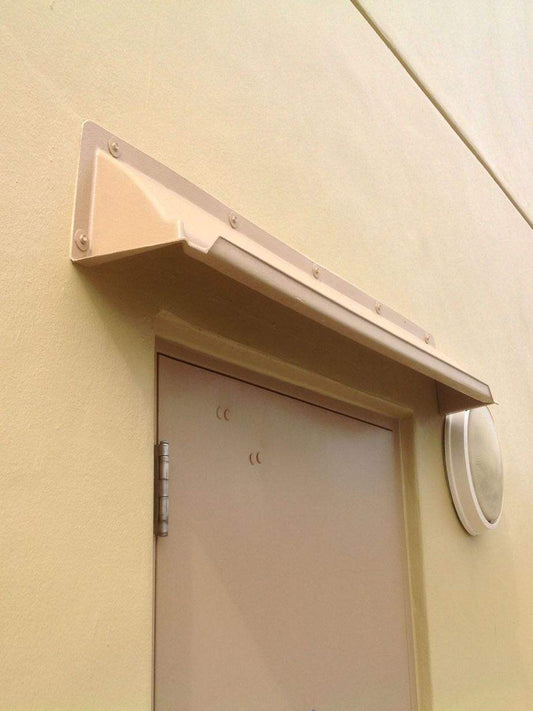 A beige door rain guards mounted above a single hollow metal door with wall mount light to the right.