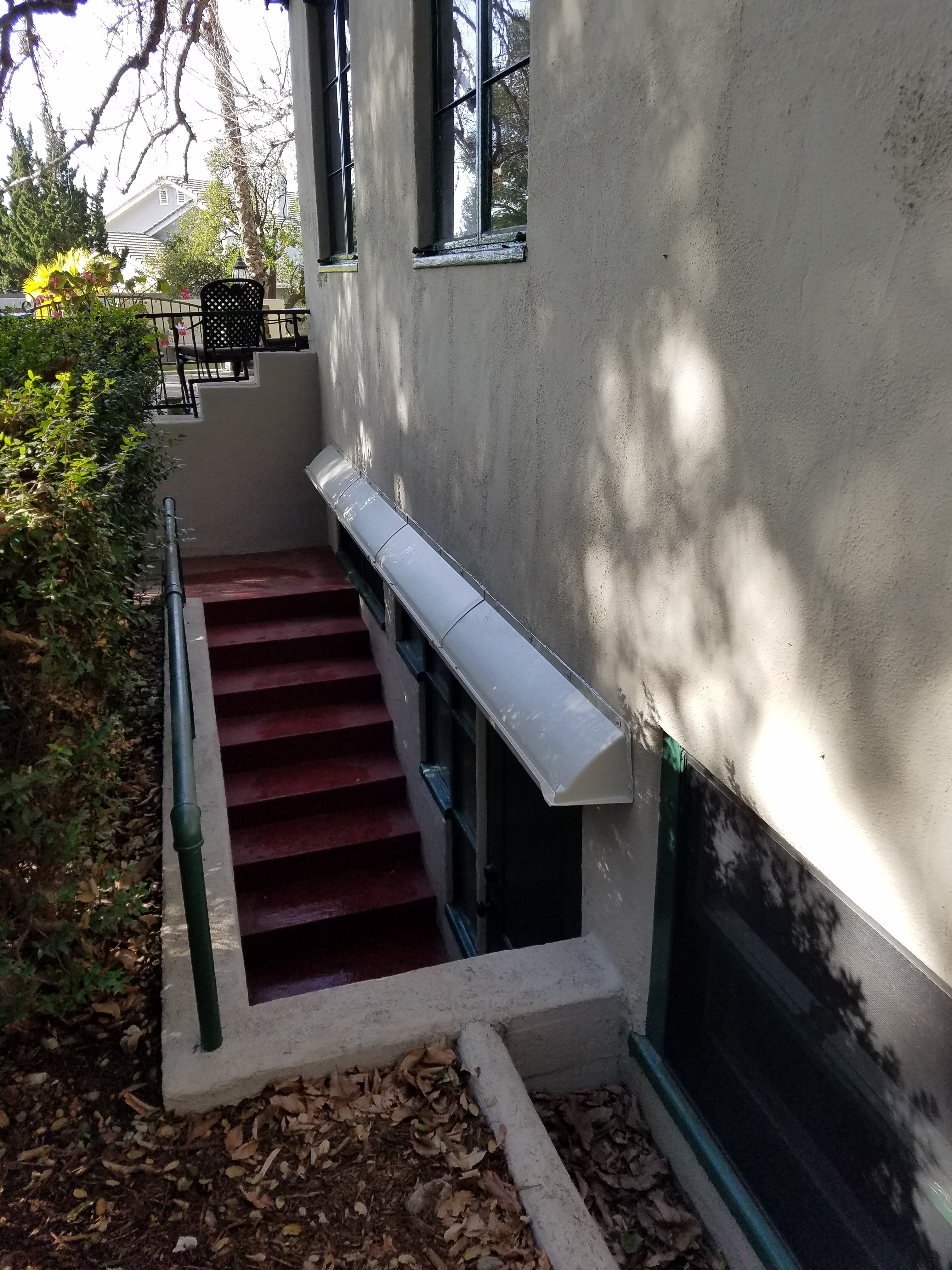 STAIRS LEADING DOWN TO LONG GRAY RAIN DIVERTER MOUNTED OVER BASEMENT WINDOW ASSEMBLY.