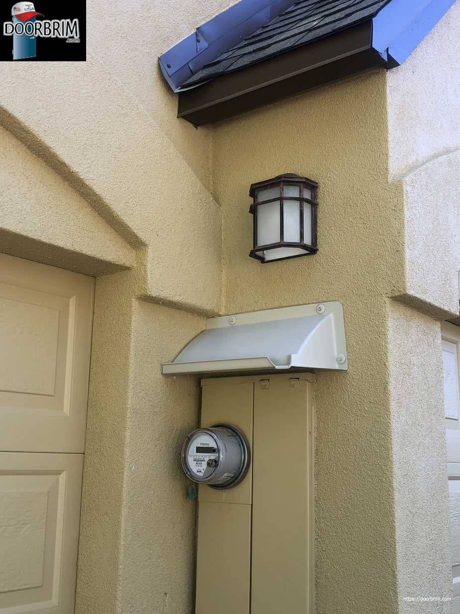 Small off white rain diverter mounted above an electric service panel next to an entrance door.